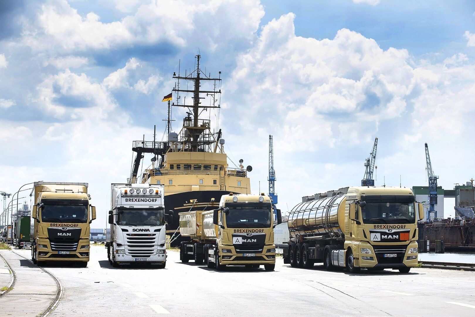 Four trucks in a port with a large ship and cranes in the background under a cloudy sky.