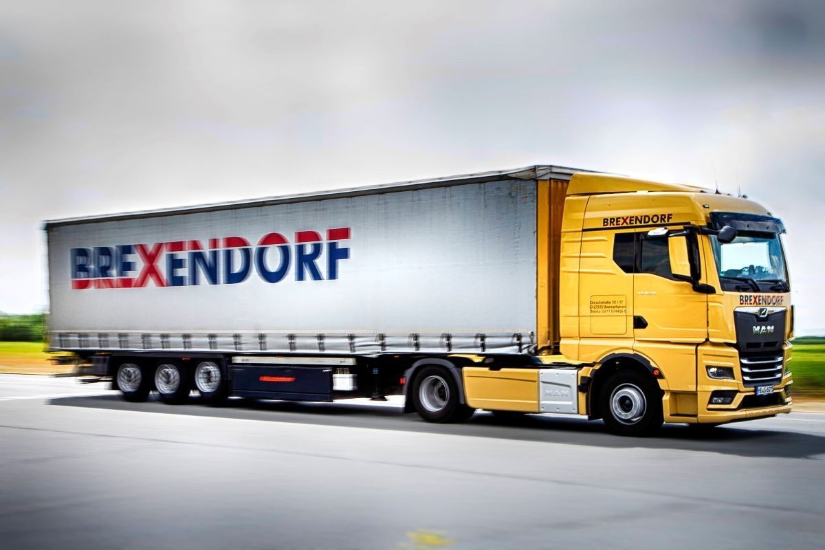 Ein gelber und weißer Sattelschlepper mit der Aufschrift "BREXENDORF" fährt auf einer Straße.