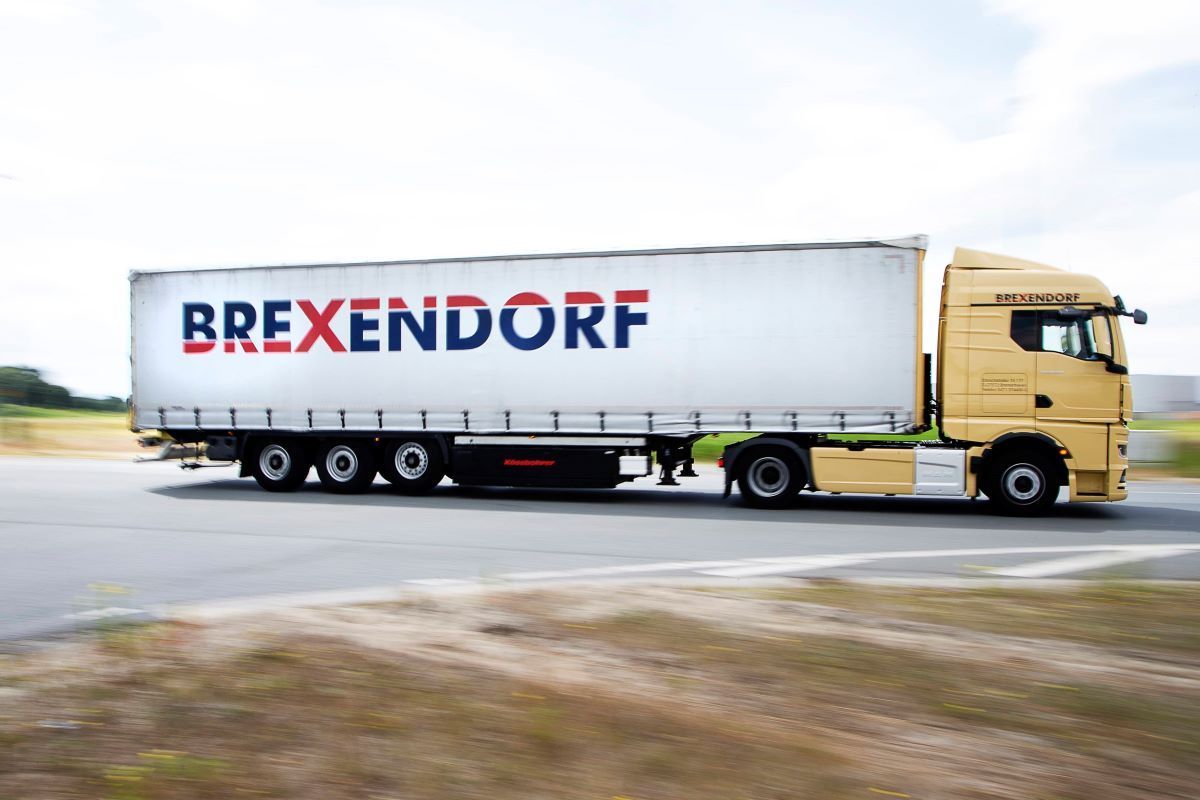 Gelber Sattelschlepper mit "BREXENDORF" auf dem Anhänger in Bewegung auf der Straße.