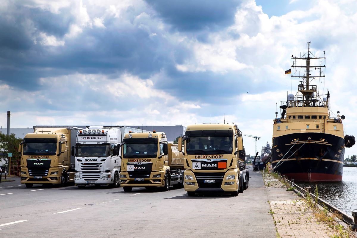 Vier in einer Reihe geparkte Lastwagen in der Nähe eines Schiffes an einer Anlegestelle unter einem bewölkten Himmel.